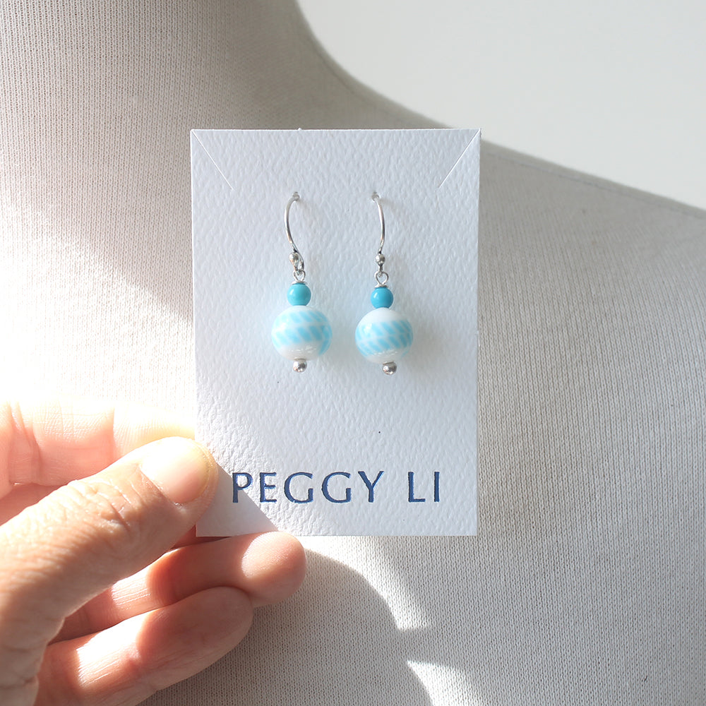 A pair of dangling earrings displayed on a white card held by a hand. The earrings have small turquoise-blue beads above larger marbled light blue and white beads, with silver hooks.