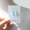 A pair of dangling earrings displayed on a white card held by a hand. The earrings have small turquoise-blue beads above larger marbled light blue and white beads, with silver hooks.