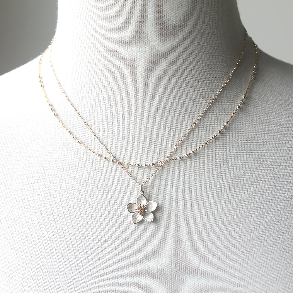 A double-strand necklace displayed on a white fabric mannequin bust. The necklace features one delicate chain with small silver beads and a second chain with a silver flower pendant that has a rose gold center.