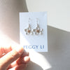 A pair of silver flower-shaped dangle earrings with gold centers, displayed on a white jewelry card printed with the brand name 'PEGGY LI'. The card is held by a hand against a textured light gray fabric background.