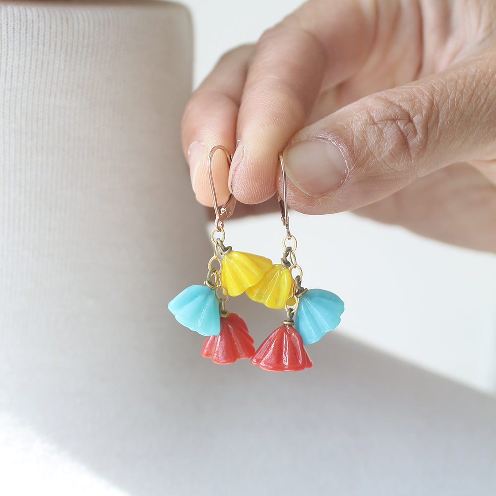 A close-up of a hand holding a pair of dangling earrings with brightly-colored blossom-shaped charms in yellow, light blue, and coral red on gold-tone hooks, against a soft white background.