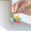 A close-up of a hand holding a pair of dangling earrings with brightly-colored blossom-shaped charms in yellow, light blue, and coral red on gold-tone hooks, against a soft white background.